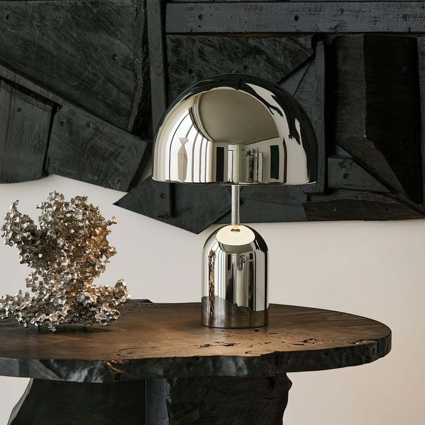 Lámpara de mesa con forma de campana cromada sobre una mesa rústica.