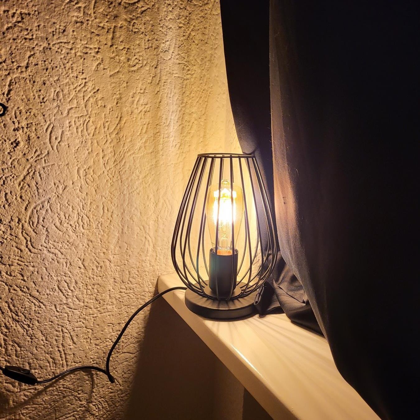 Lámpara de jaula negra iluminada sobre una repisa con pared texturizada.